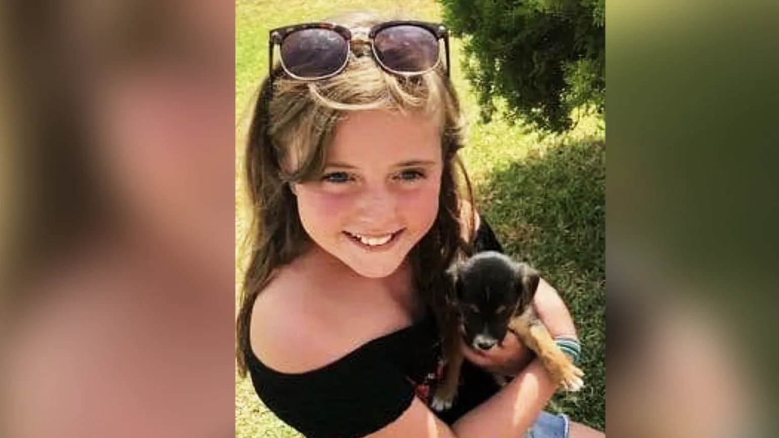 Nia Day Nia, aged about nine. She wears a black off the shoulder top and shorts and has sunglasses on her head. She has long light brown hair. She is holding a black and tan puppy. She is smiling straight ahead but the photo is shot from above so not towards the camera.