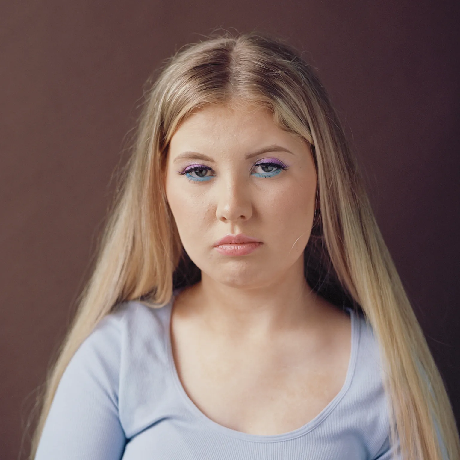 Museum Wales Head and shoulders shot of a girl with long blonde hair. She wears a scoop neck light blue cotton top and has striking eye make up on, with electric blue eye liner underneath and light purple eyeshadow on her lids. She looks at the camera without smiling.