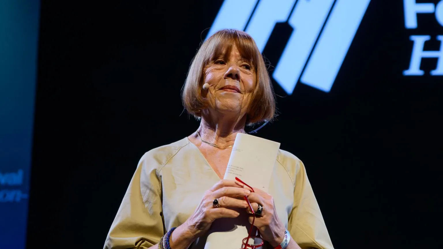 Gisele on stage at the hay festival. she is wearing a loose fitting, v neck beige top and has red hair in bob style. she is clasping her hands in front of her chest, holding a folded piece or paper and red rimmed glasses.