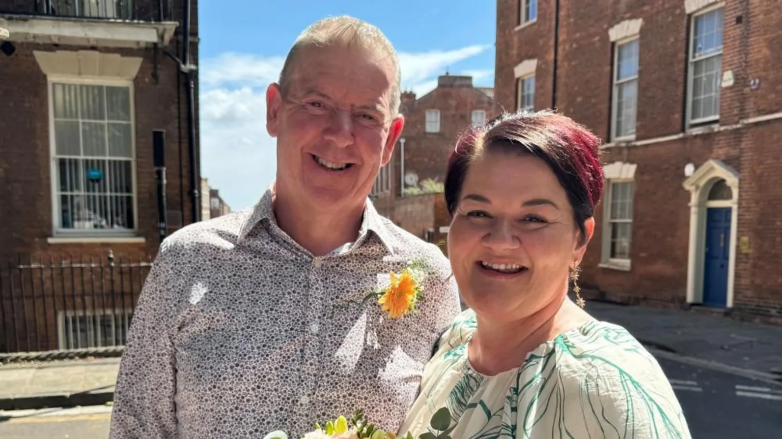 Emma is stood next to her husband John. She is holiding flowers and has short dark hair with a red highlight. He is wearing a patterned shirt with a flower stuck on.