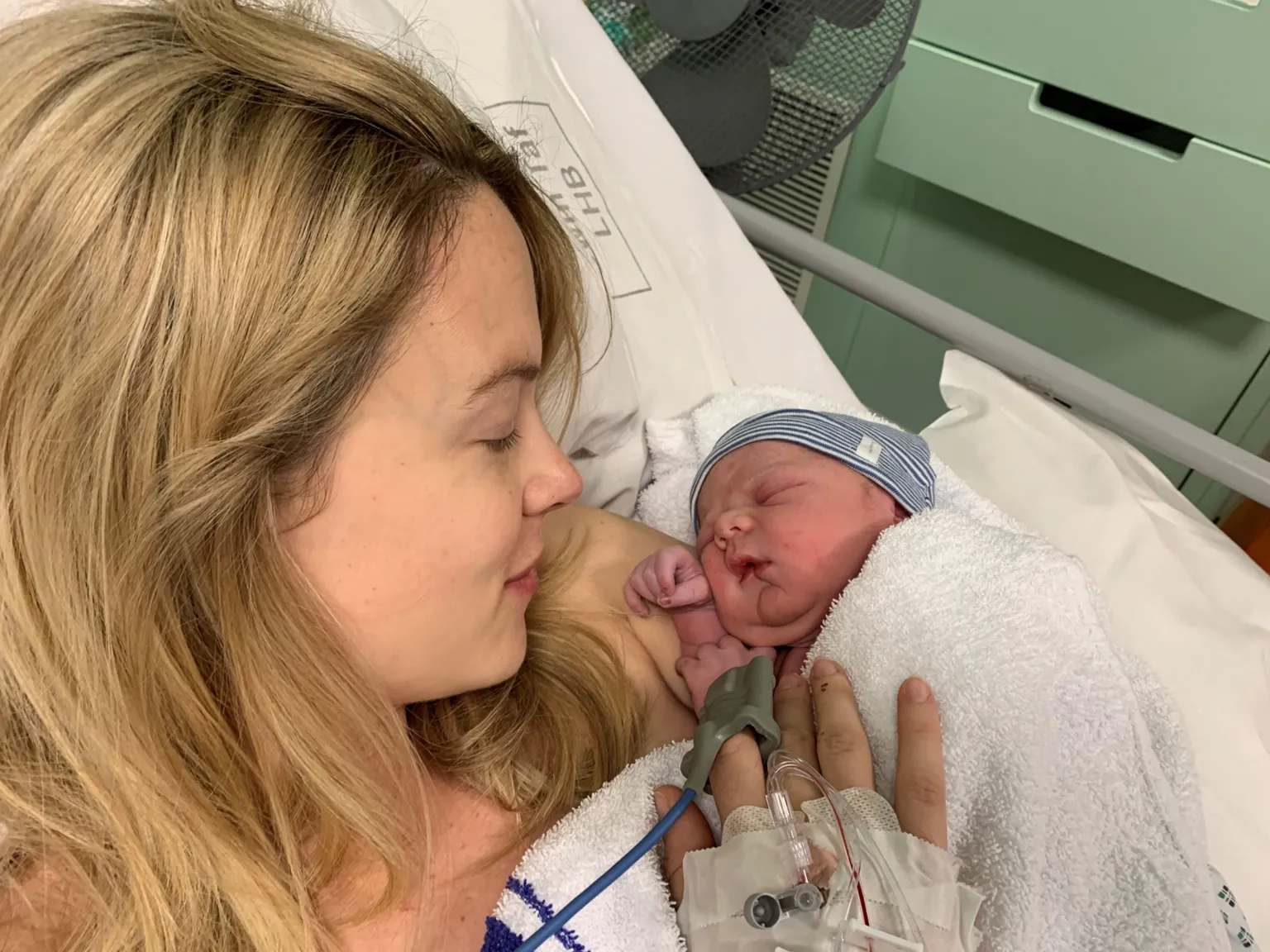 Kimberley Nixon A blonde woman lying in a hospital bed with tubes coming out of her hand. She is looking down at a tiny baby with a blue and white striped hat on in her arms.