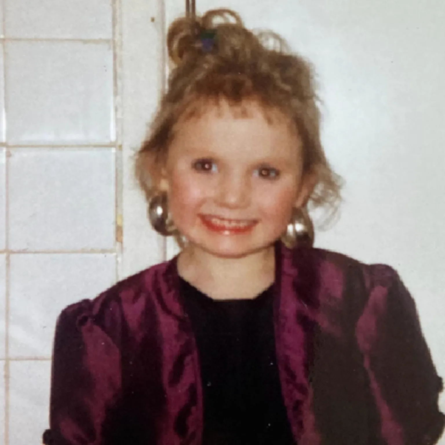 Michaela Allen Seven-year-old Michaela smiles at the camera, she has her light brown hair tied up and is wearing huge silver earrings. She is wearing a plum satin jacket with shoulder pads and is stood in front of a white wall.