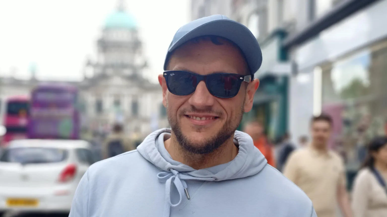 A man, with brown stubble, wearing a blue cap, blue hoodie and sunglasses.