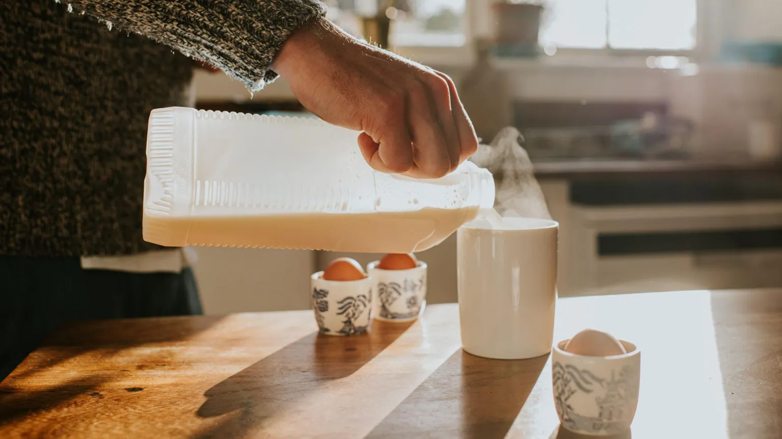  A man's hand pouring milk into a steaming mug with boiled eggs on the wooden worktop