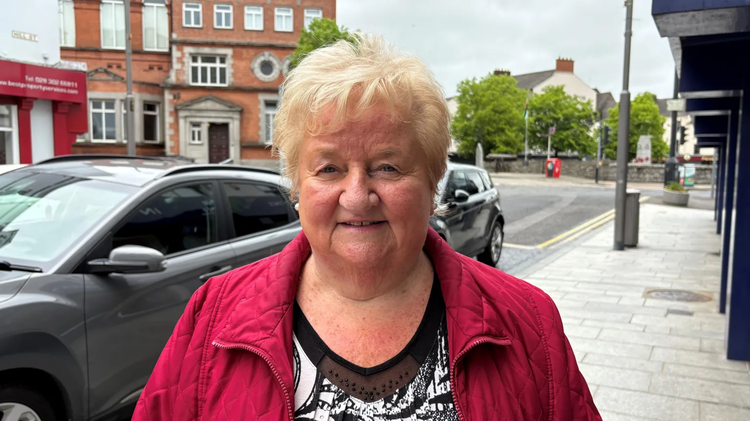 Marie Byrne, a woman with short blonde hair, smiles at the camera while standing on Hill Street in Newry. She is wearing a red quilted jacket over a black and white patterned top. Shop fronts and a tall red brick building - Newry Town Hall - are in the background. There are cars parked along the pavement.