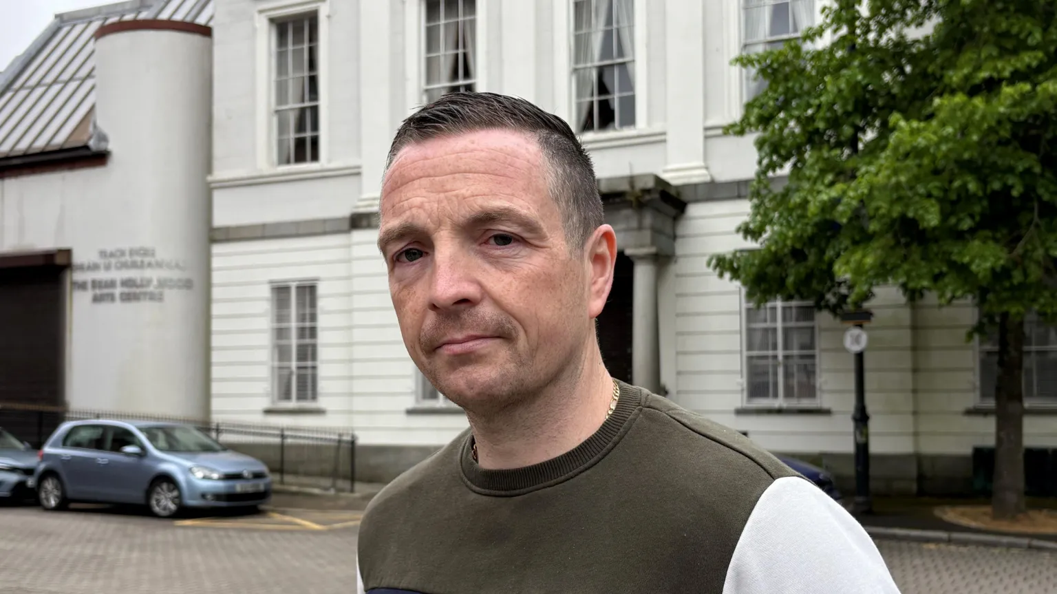 Patrick Smith, a man with short dark hair, stands in front of the former Sean Hollywood Arts Centre in the centre of Newry. He is wearing an olive green sweatshirt with cream sleeves. There is a tree in front of the building and two blue cars parked outside. 