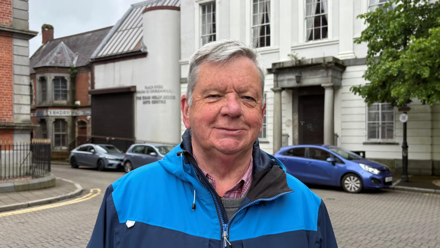 Ray Casey, a man with short grey hair, smiles at the camera while standing in front of the former Sean Hollywood Arts Centre. He is wearing a two-tone blue rain jacket over a grey jumper and red checked shirt. 