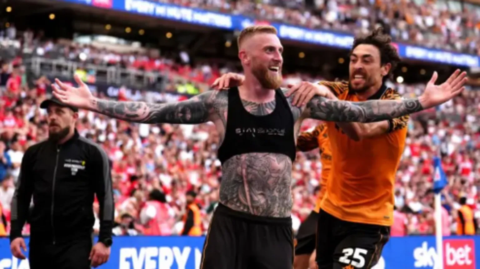  A footballer, with his top off exposing a sports vest, holds both hands up in celebration as a team-mate reaches out to him. Behind them fans in the terraces go wild.