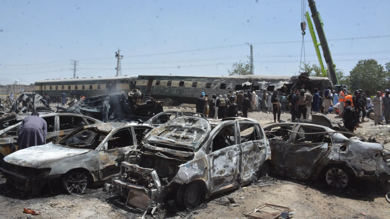 Burnt-out cars are seen in front of a derailed train, while people stand around.