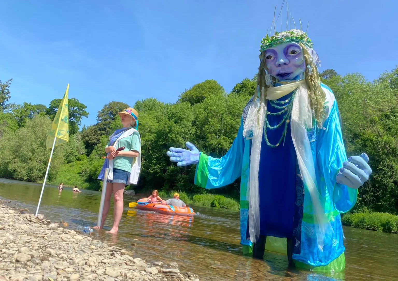 A large puppet covered in blue and green robes stands in the river. Behind it is a dingy on the water.