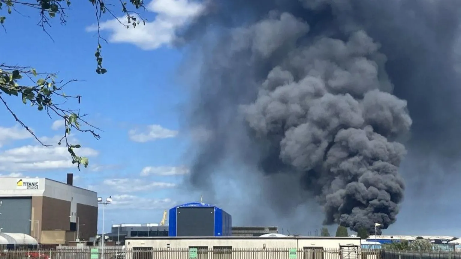 Large plume of grey smoke against a blue sky with white clouds in east Belfast. The Titanic Studios building is to the left and there is a blue building in the centre.