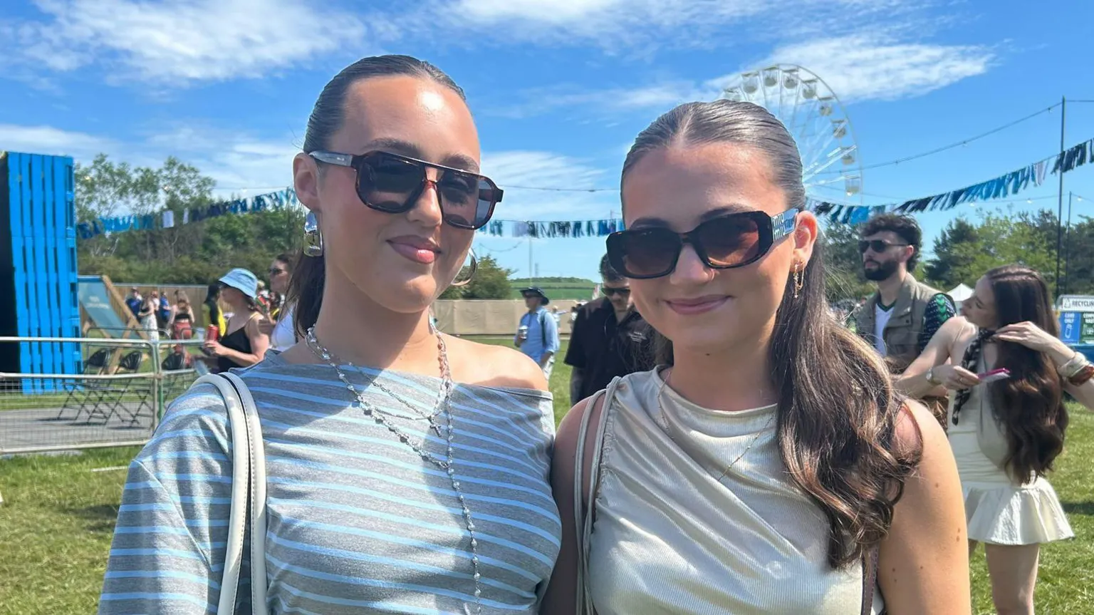 Two young women in sunglasses smile at the camera on a music festival site.