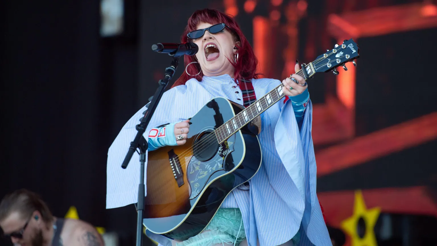 CMAT sings, wearing black sunglasses and a blue cape, her hair red, into a microphone while holding a guitar.