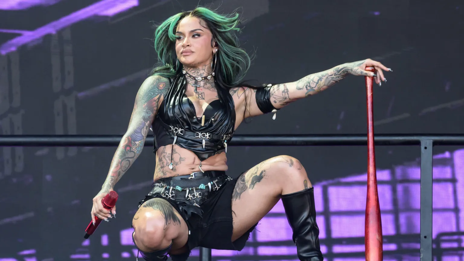 Getty Kehlani performs on stage wearing a black leather bra top and shorts, paired with black knee-high boots. She has black hair with green highlights. She is crouched down on the stage, leaning on a red baseball bat.