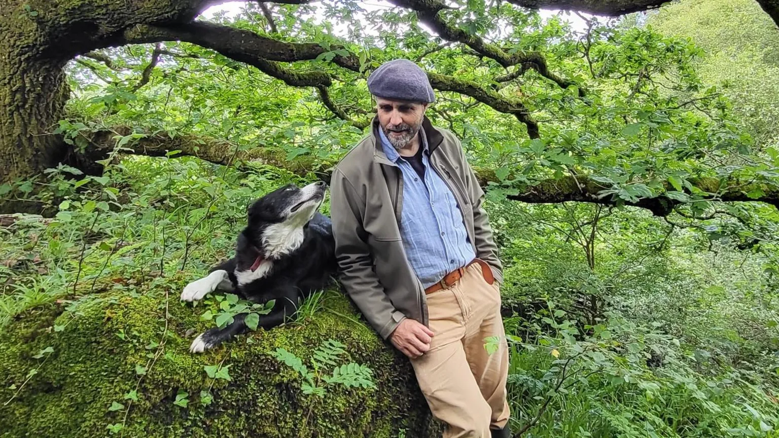 Eoghan Daltun A man wearing a grey cap, beige trousers, a blue shirt and a grey jacket is photographed standing beside a dog which is a collie breed. The dog is looking up at him and they are leaning on a tree in his temperate rainforest. Greenery everywhere. 