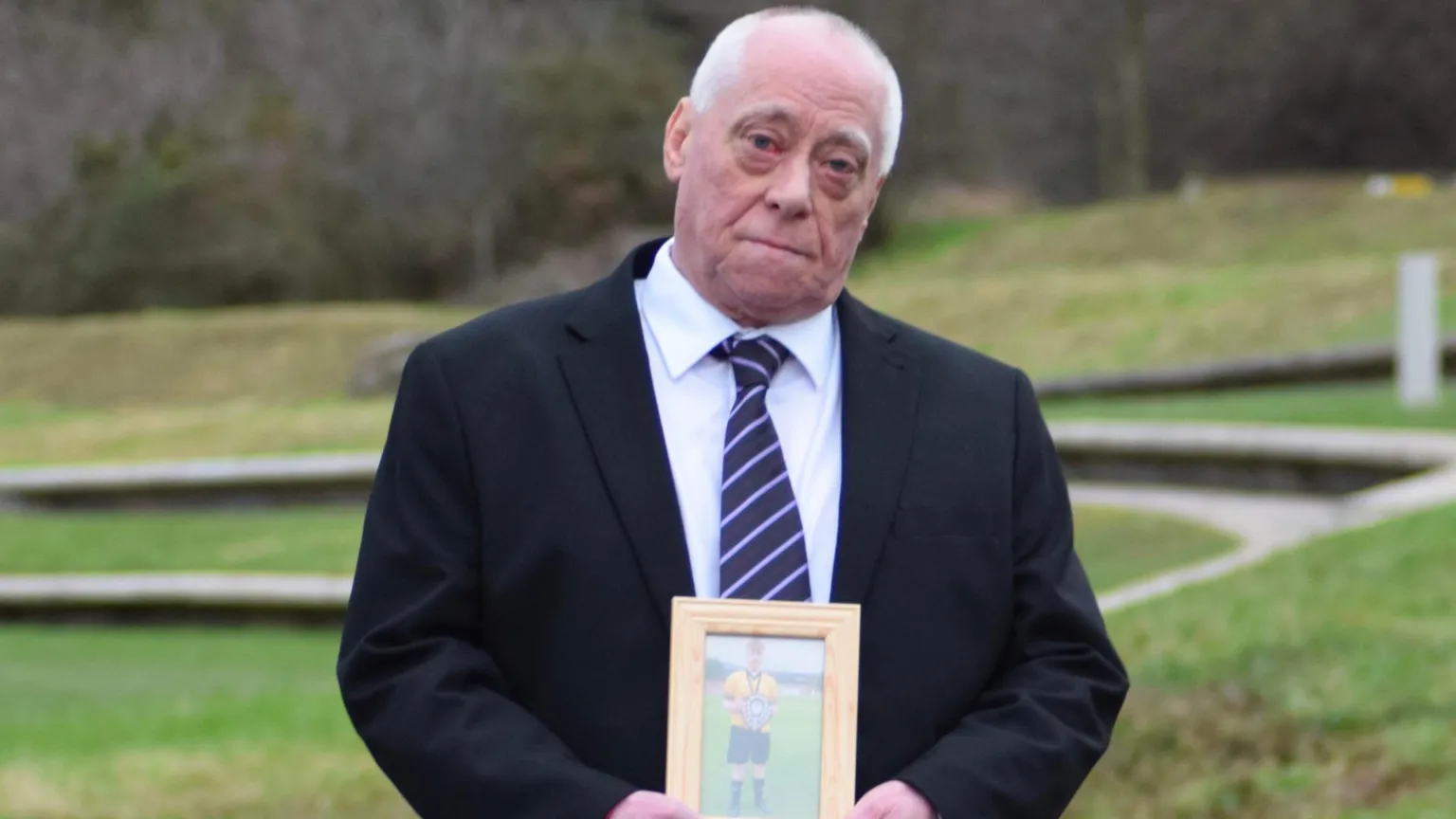 Alan Knox stands outside the Scottish Parliament wearing a suit with grass behind him holding a wooden photo frame with a picture of his son Lewis Knox inside.