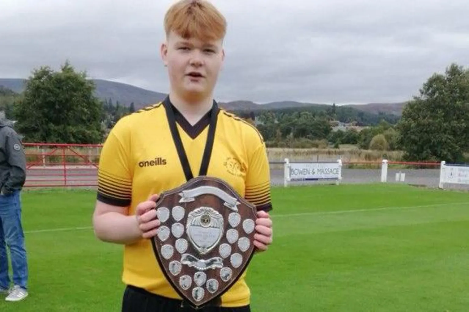 Liz Knox Lewis Knox who played for Fort William Shinty Club in his yellow and black strip holding a wooden trophy covered in small silver shields.