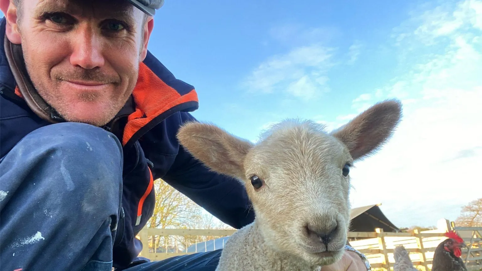 Mark Rutherford A man in a flat cap and outdoor working clothes, knelt down smiling into the camera. Next to him is a sheep, and behind is a farm.