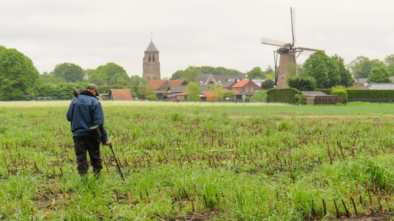 Filip Krapels Filip with his back to the camera is wearing a blue jacket and black trousers, with a spade slung over his left shoulder. He scans the ground with a metal detector, the bottom of which is obscured by the long grass. In the background there is a windmill and a small village with a church spire.