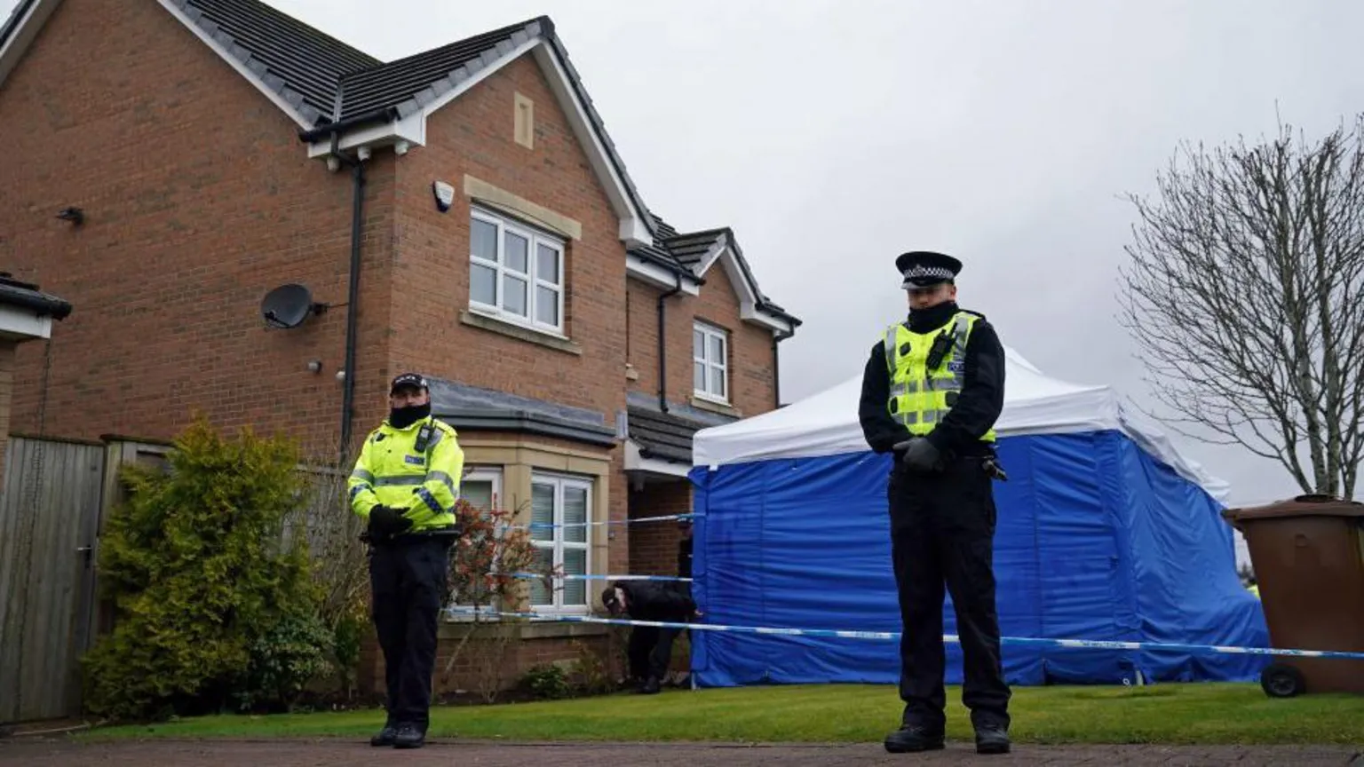  Two police officers, wrapped up against the cold, standing outside a detached, two-storey modern house. A square blue tent with a white roof is pitched on the front lawn of the house and there is police tape preventing people from entering the garden. 