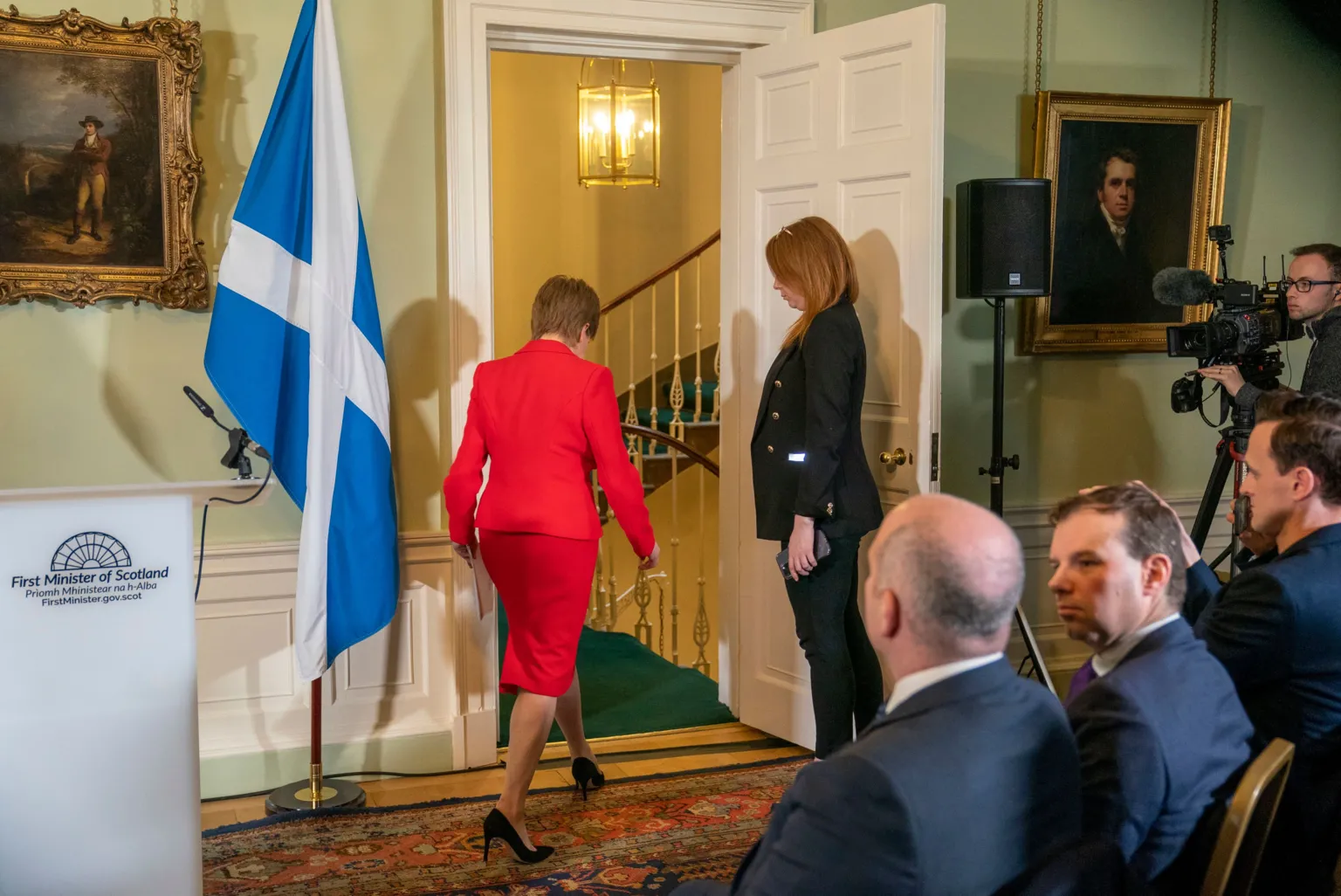  Nicola Sturgeon in a red suit walking out of a room full of journalists. Glenn Campbell and James Cook can been seen seated in the foreground. 