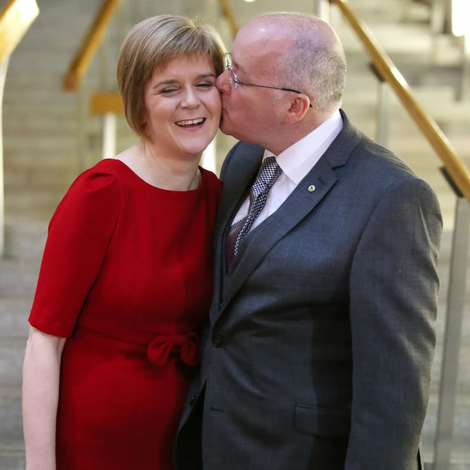  Nicola Sturgeon and Peter Murrell at the bottom of the staircase in the Scottish Parliament building. Sturgeon is wearing a red dress; Murrell is in a suit. He is kissing her on the cheek and she is smiling. 