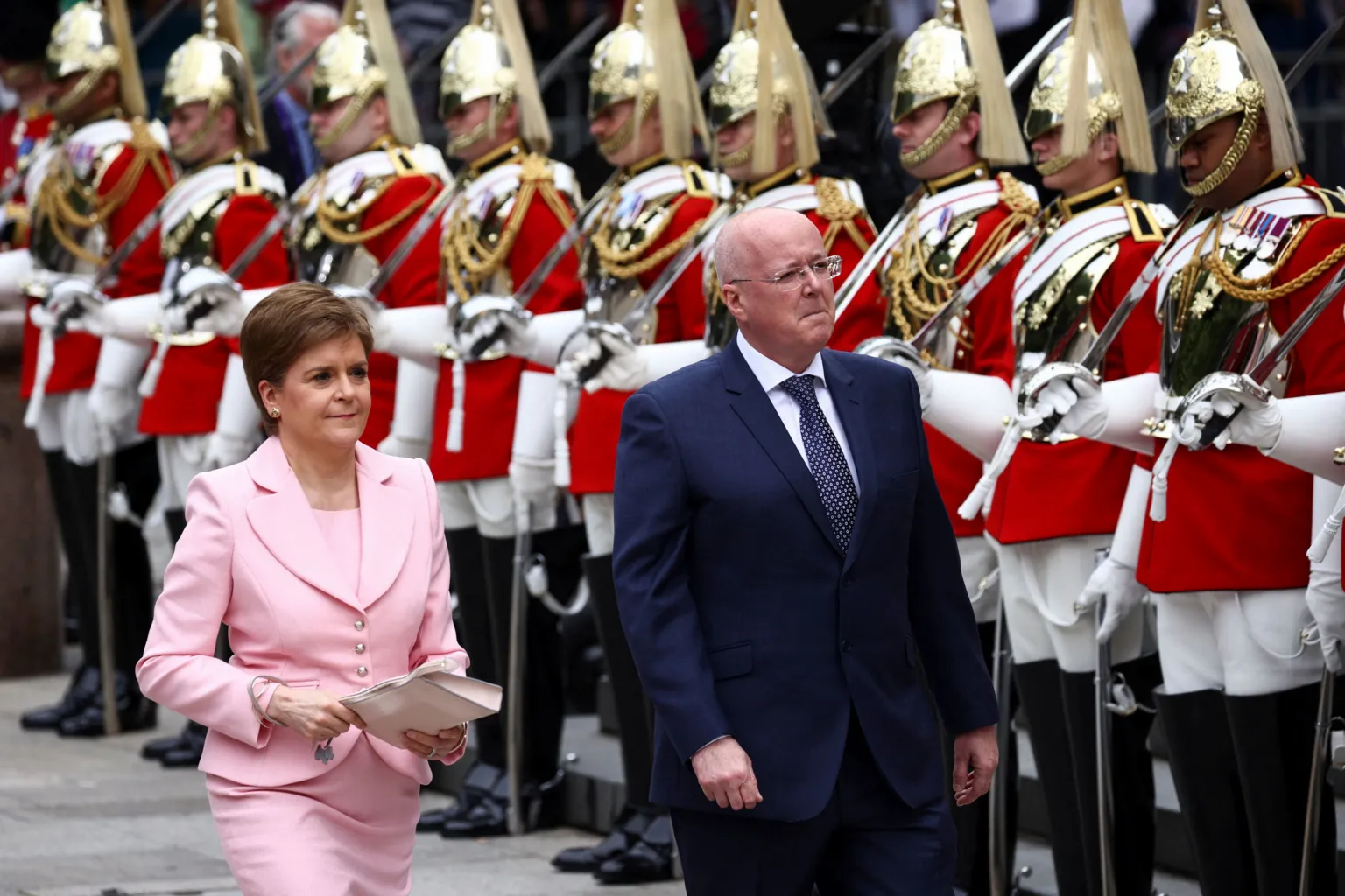  Nicola Sturgeon and Peter Murrell walk in front of a line of servicemen dressed in elaborate regalia. Sturgeon is wearing a pink suit. Murrell is in a navy blue suit and tie. 