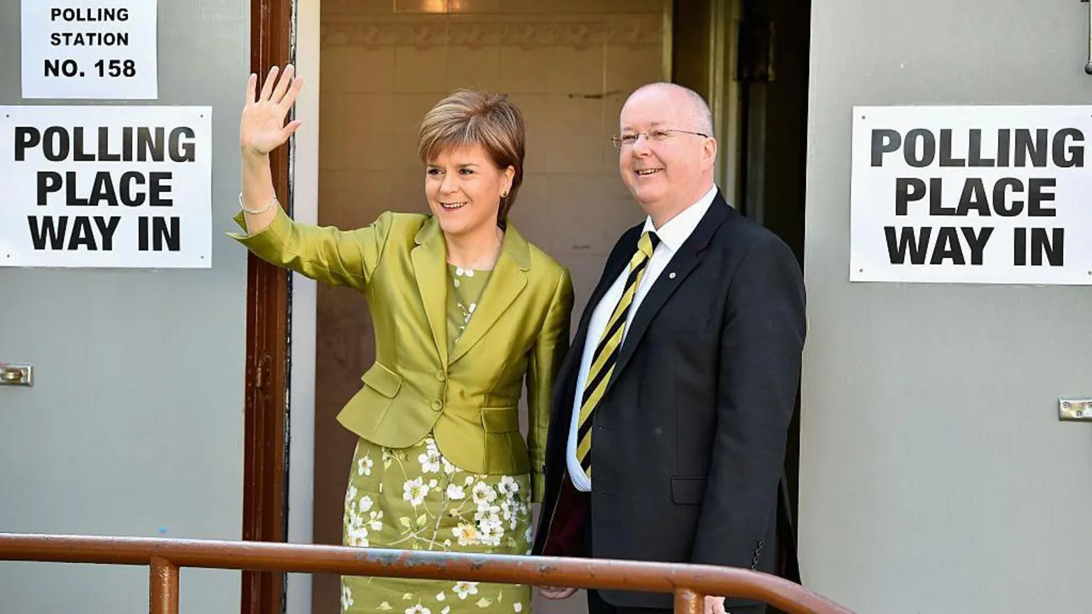  Nicola Sturgeon in a green floral dress and matching cropped jacket. She is standing next to Peter Murrell, dressed in a dark suit, white shirt and black and yellow striped tie. They are standing outside a polling place on election day, posing for the cameras.