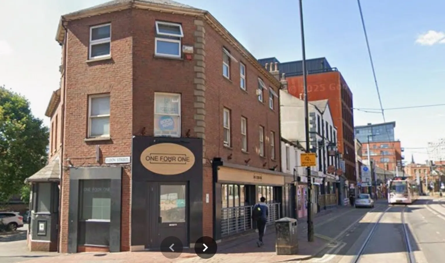 Google In the foreground is a three-storey red-brick building with a gold coloured oval-shaped sign bearing the words One Four One above a black doorway. Further down the city centre street are a mixture of old and new shops and offices and a tram line on the road. It is a sunny day with a blue sky.