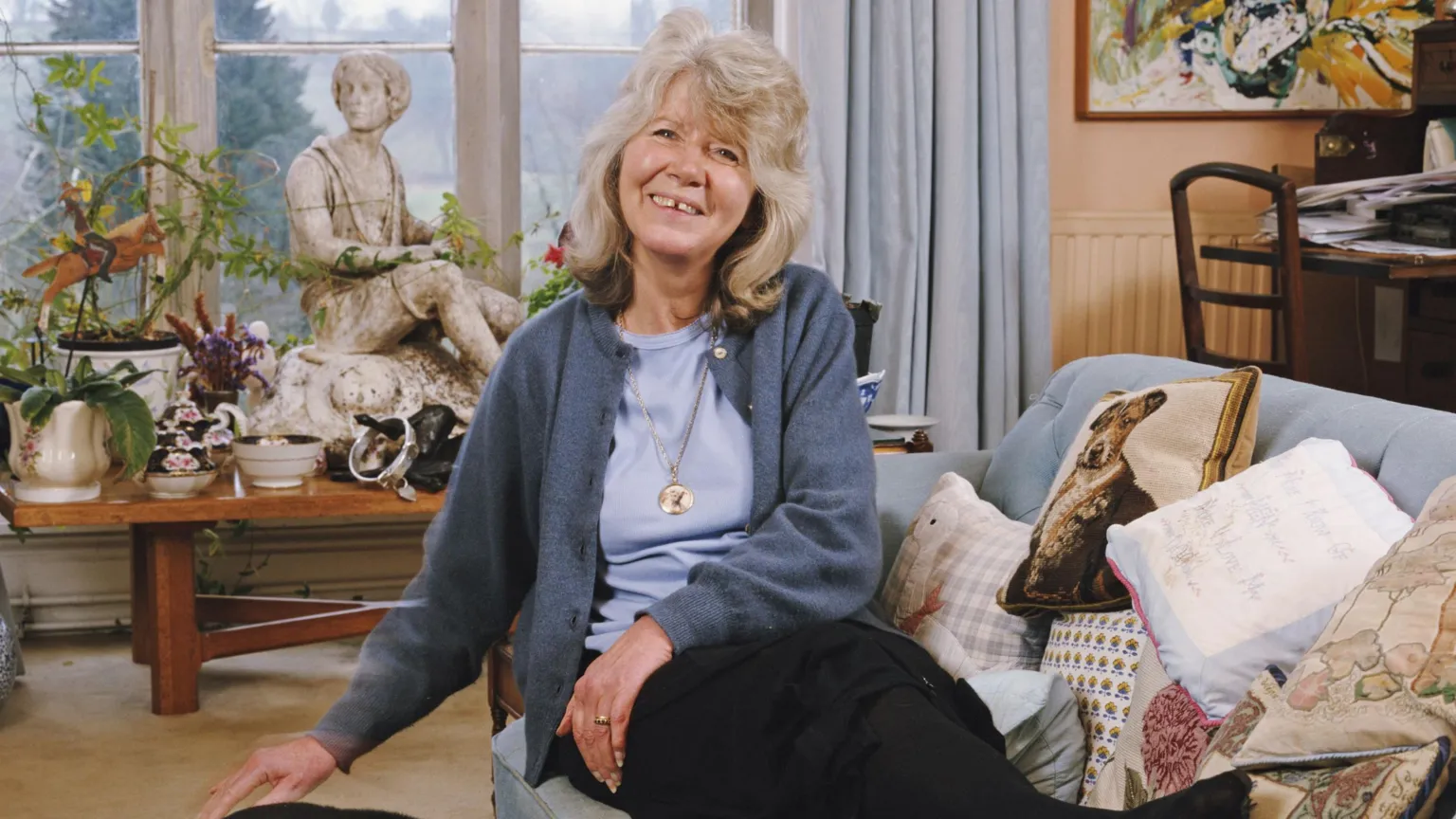 Dame Jilly Cooper in 2000. She's sat on a sofa in a living room full of plants and antiques. 