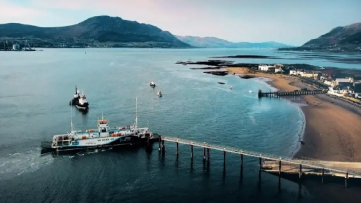 An aerial view of the Carlingford Lough Ferry coming into dock at Greencastle, County Down. A long pier stretches into the water in front of a long, sandy shore. Houses and small buildings line the beach. There are mountains hugging the coastline around either side of the lough.