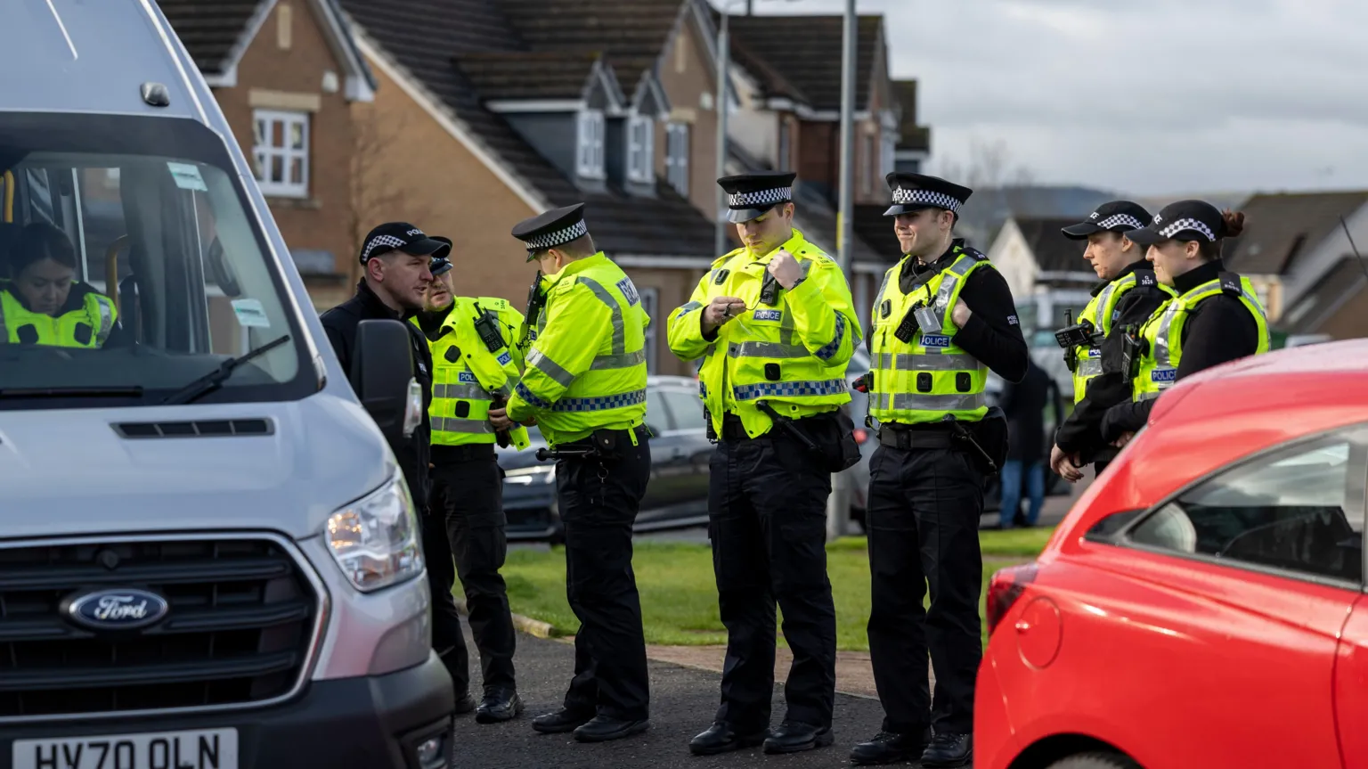 Seven police officers in high viz vests in a residential area, standing in a line next to a white police vehicle with another officer sitting inside.