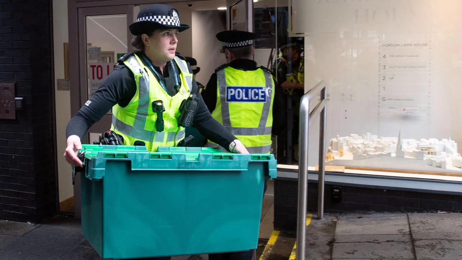 A female police officer carries a large green plastic box out of the SNP's headquarters in Edinburgh following the arrest of Peter Murrell in April 2023. Another officer is heading back into the building, which has a frosted glass exterior.