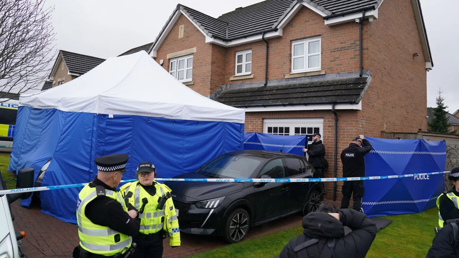 A blue and white police tent outside a brick house, next to a black car. Two police officers in a high viz vests are at the left of the image, and other officers in black clothes are at the side of the house.