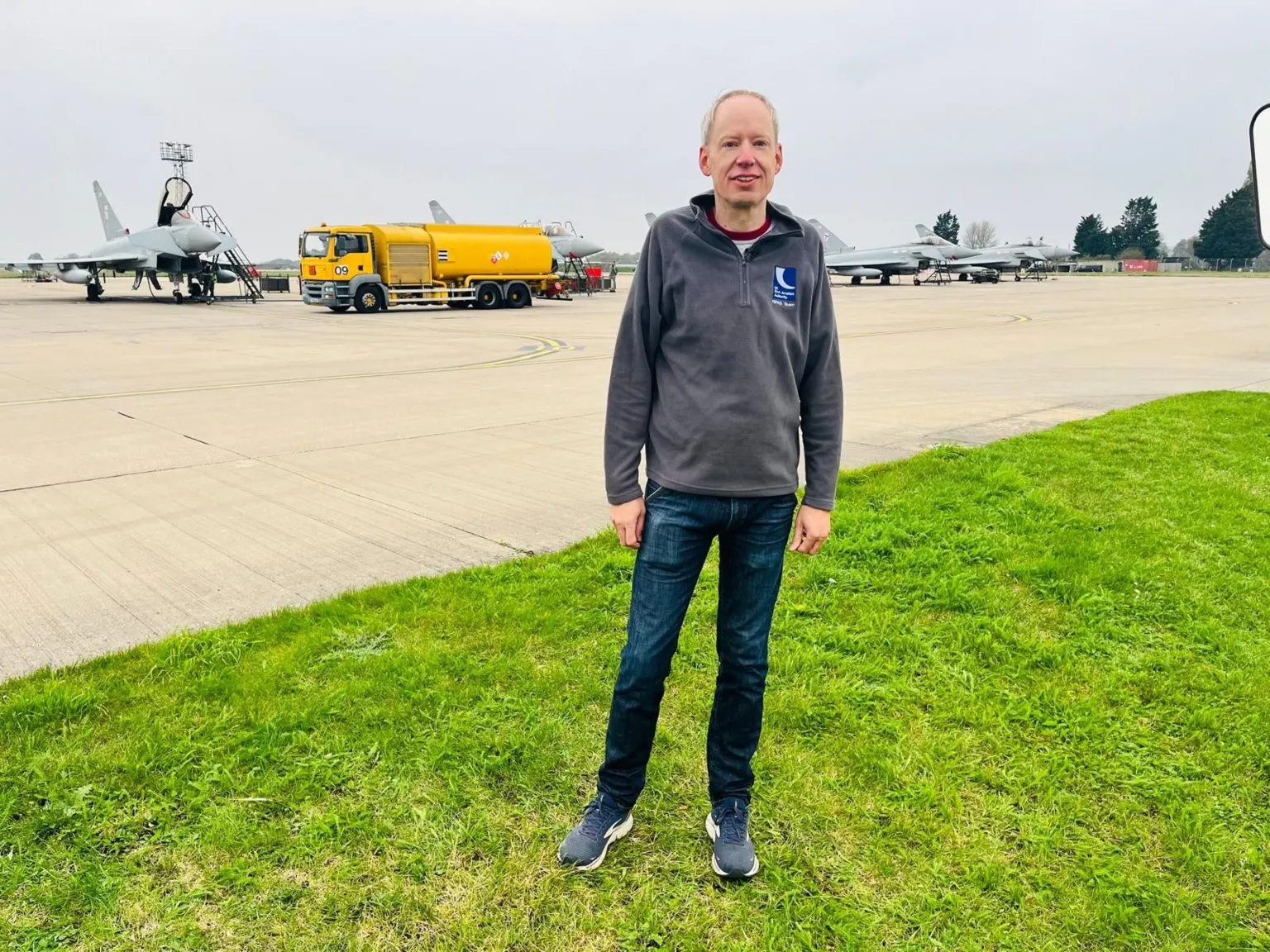 CAA A man standing on grass in front of a fighter jet on an airfield.