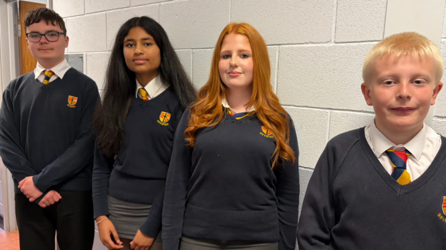 Four pupils - two boys and two girls - in a row next to a wall wearing navy school jumpers with a logo, white shirts and colourful ties.
