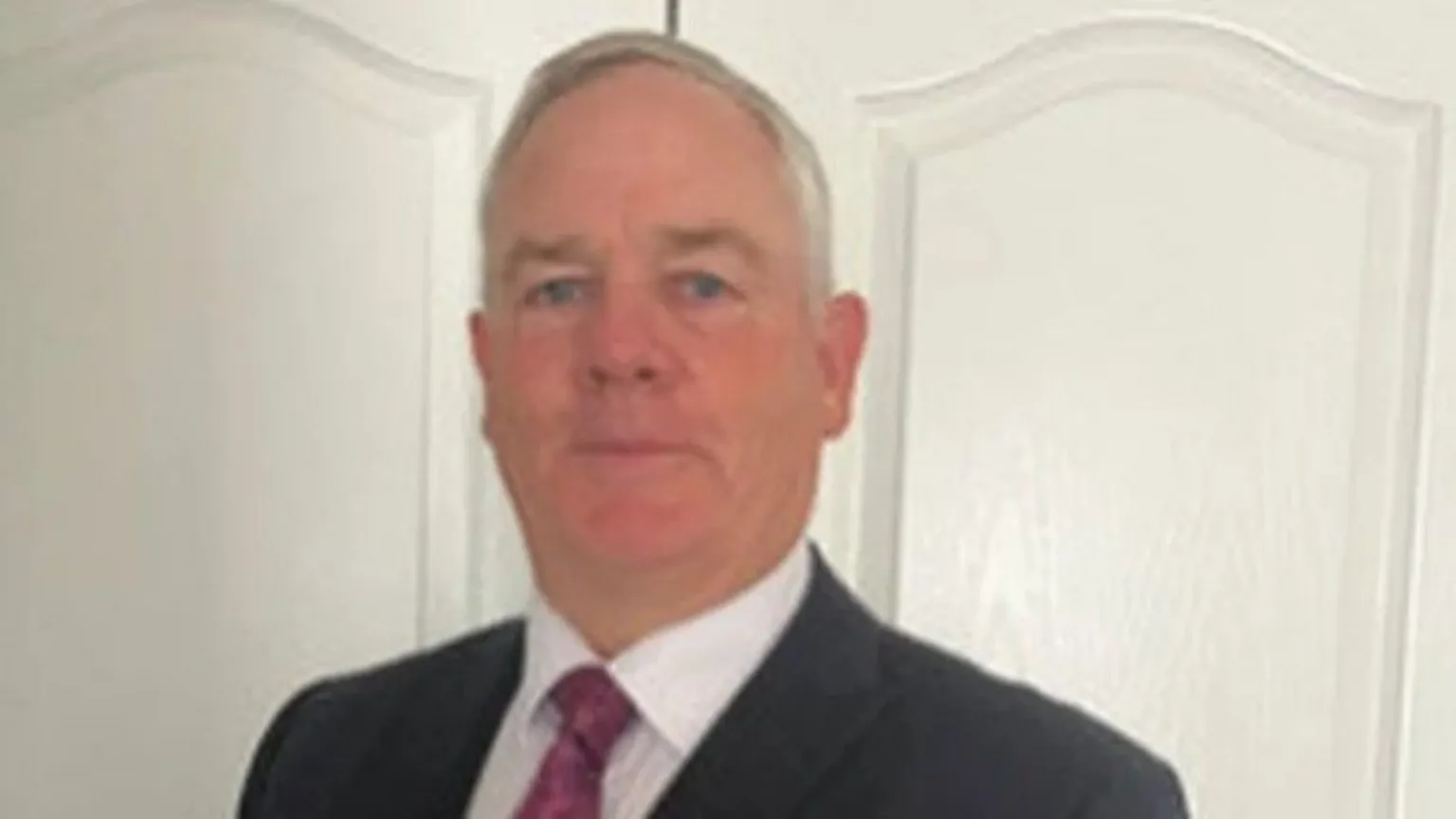 ICLVR Eamonn Henry, a man with grey hair and blue eyes, looks directly at the camera. He is wearing a dark suit, a white shirt and a red tie. He is standing in front of a white cupboards.