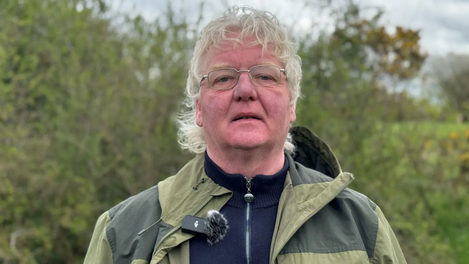 James Orr has white shaggy hair and glasses. He is wearing a light green waterproof coat over a dark blue quarter zip top. He is standing in front of a green hedge.