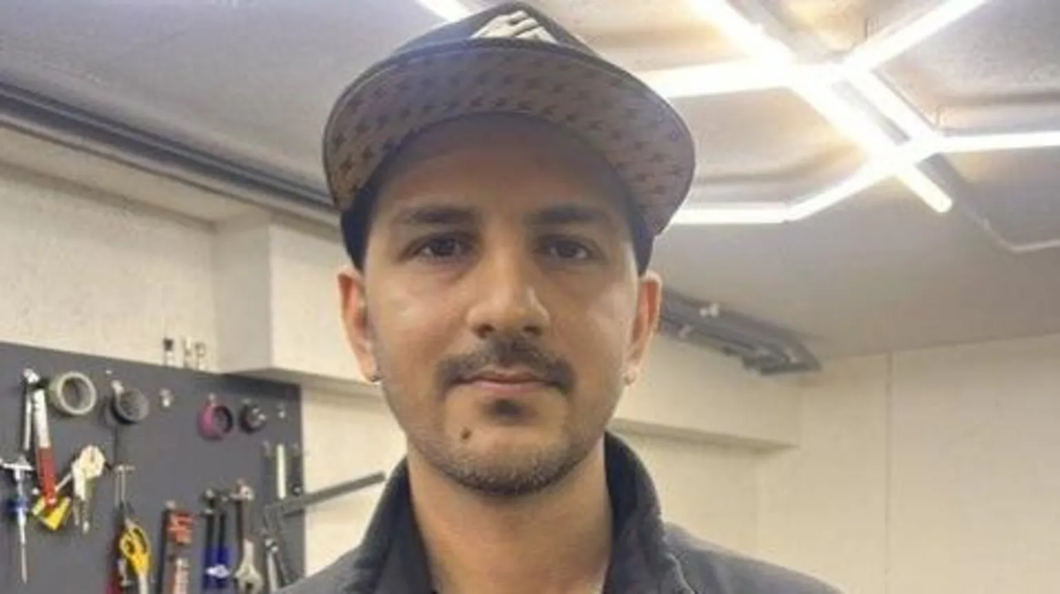 Head and shoulders image of Saim Shahzad in his e-bike repair shop in Aberdeen. He is wearing a brown and cream baseball cap and a grey zip. He has a short dark mustache and short drak/grey goatie-style beard. On the wall behind is right shoulder the is key rack with lots of sets of keys.