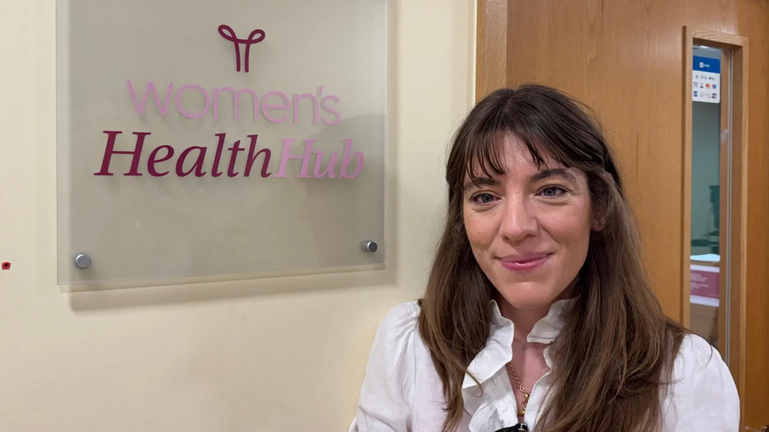 Dr Jessica Langtree-Marsh is smiling at the camera in a heads-and-shoulders photo. She has long brown hair and is wearing a white top. She is standing next to a sign in a corridor that says 