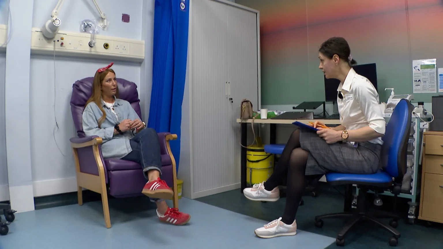 Cancer patient Jemma Sleeman is sitting in a medical consultation room chair. She has long long brown hair and is wearing a blue shirt over a white T-shirt, jeans and red trainers. She also has a red hair band. She sits opposite oncology research doctor Madalina Chifu, who has shorter dark hair tied back in a bun. Dr Chifu is also wearing a blouse, skirt and trainers, plus is holding a clipboard to make notes during the appointment.