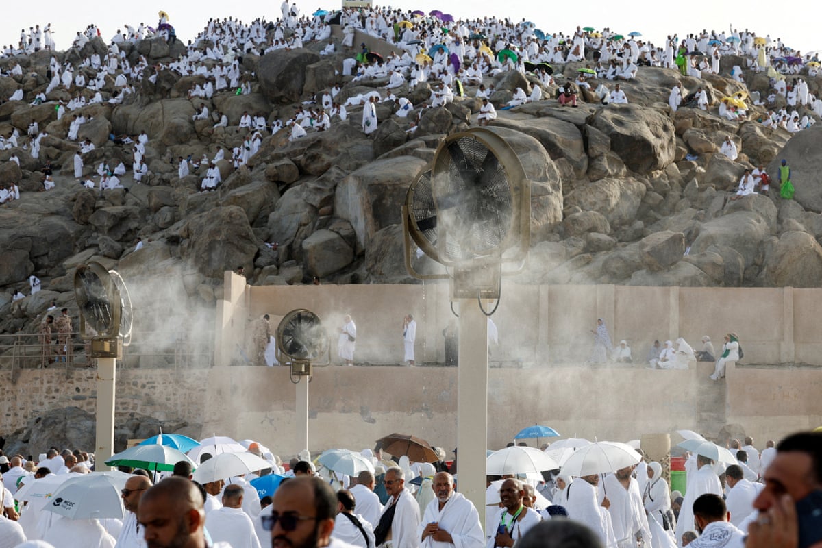 A crowd of pilgrims in white robes gather by misting fans to beat the heat, with mount Arafat in the background.