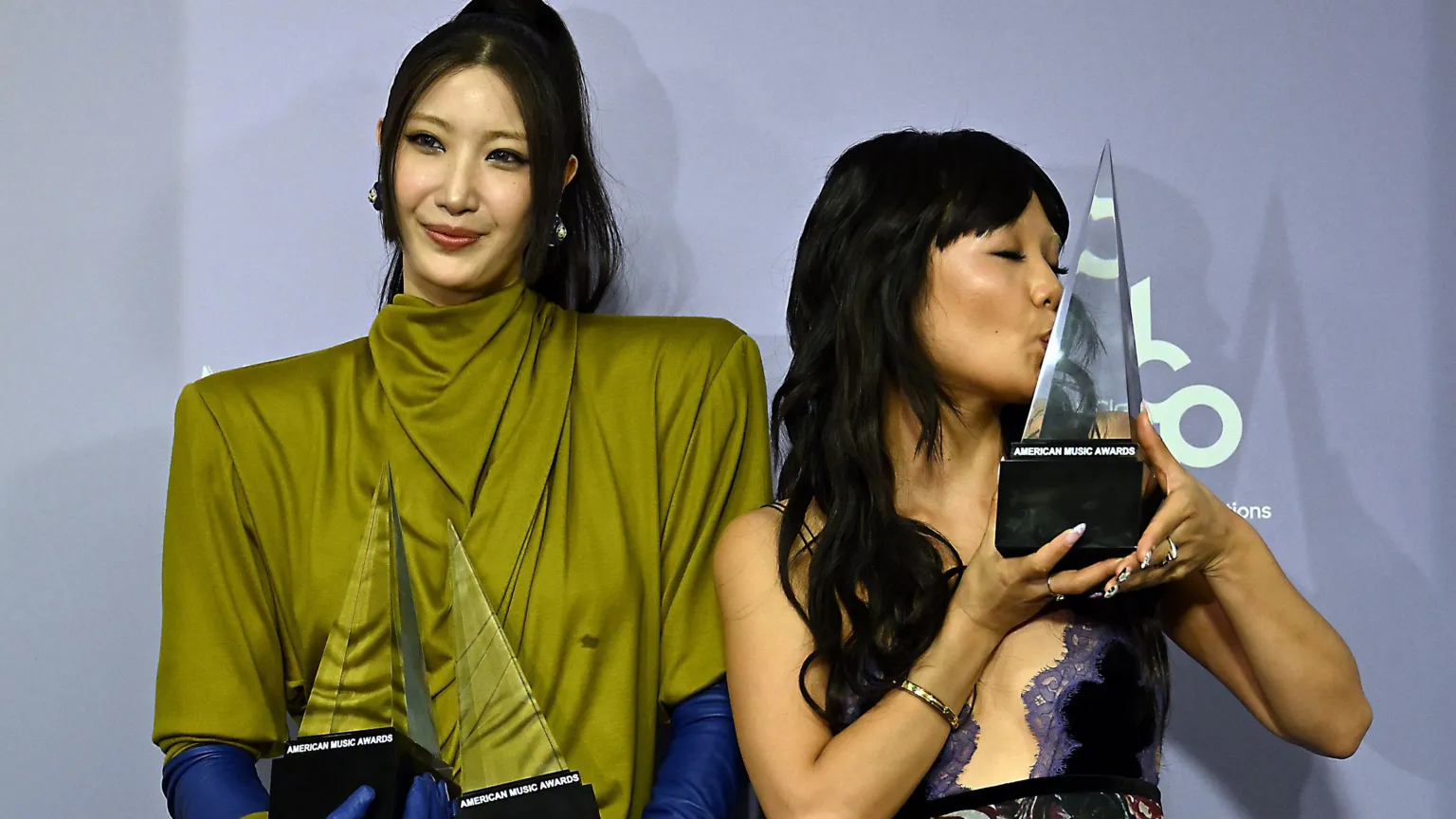  EJAE, wearing an olive green top, and Rei Ami, wearing a purple and black lace dress, hold their awards backstage.