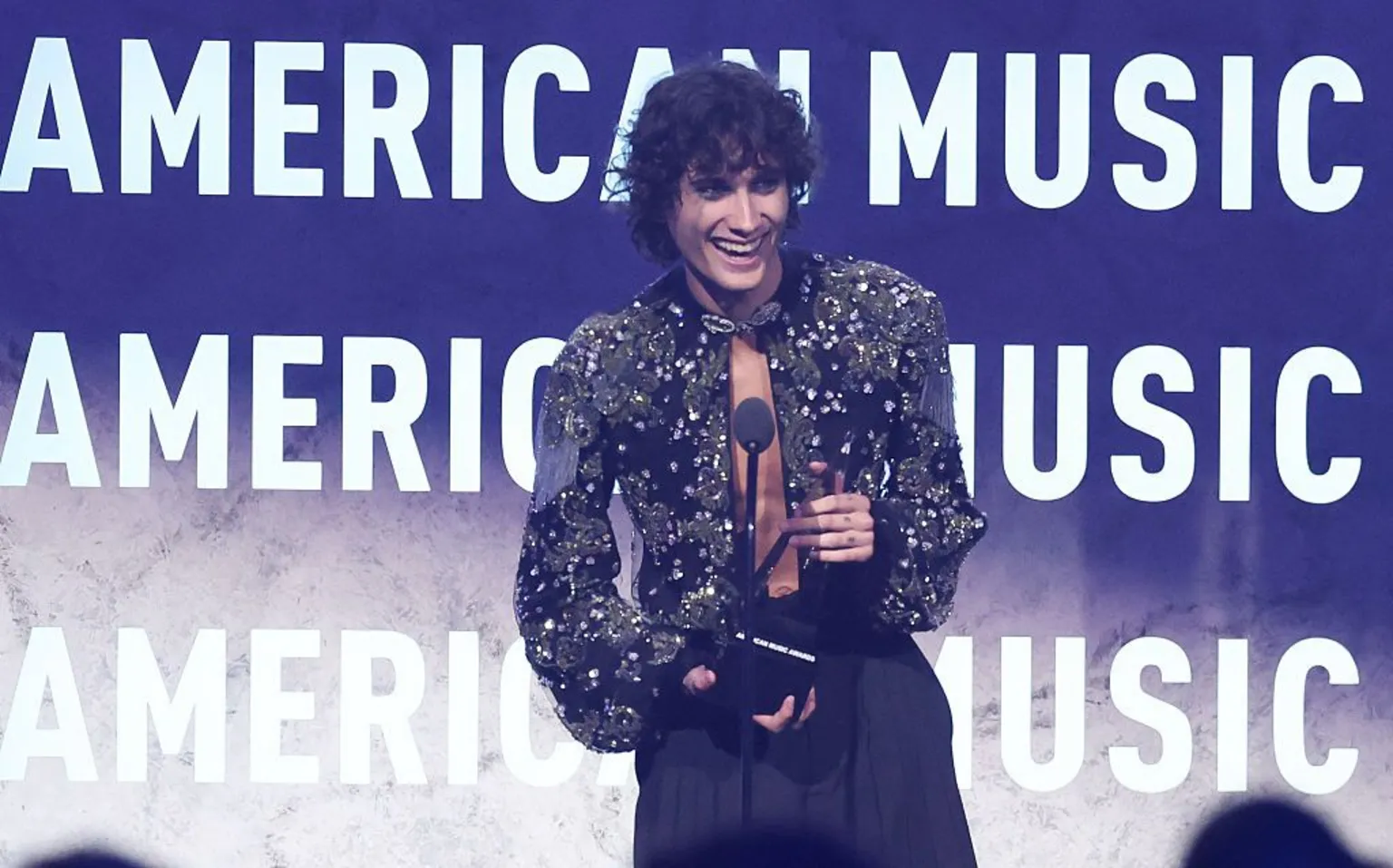  Sombr collecting an award on stage - he is wearing black trousers and a dark glittery top slashed down the middle at the front.