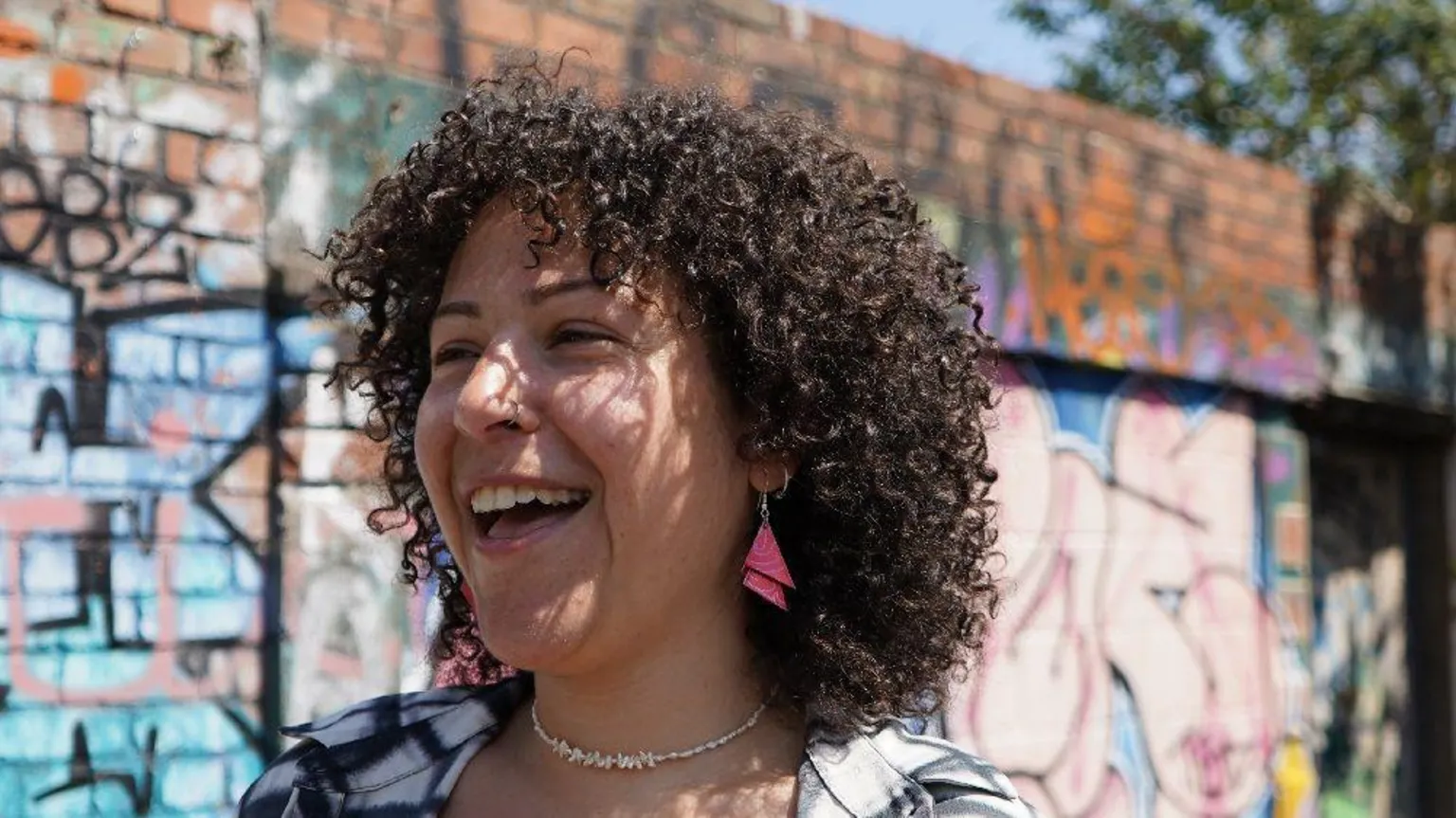 Handout Engy Elboreini smiles at the camera. She has curly dark-brown hair and pink earrings. She is standing in front of a wall spray-painted with graffiti.