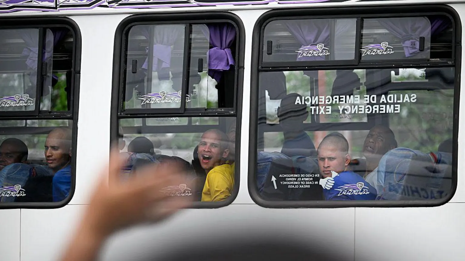 Federico PARRA / AFP via Inmates look through the window of a bus as they are transferred from the Internado Judicial Barinas (Injuba) prison, in Barinas, Venezuela, on May 25, 2026. Their heads are shaven and they are wearing prison T-shirts, some of them blue, others yellow. One inmate appears to be shouting something to relatives gathered outside the prison.