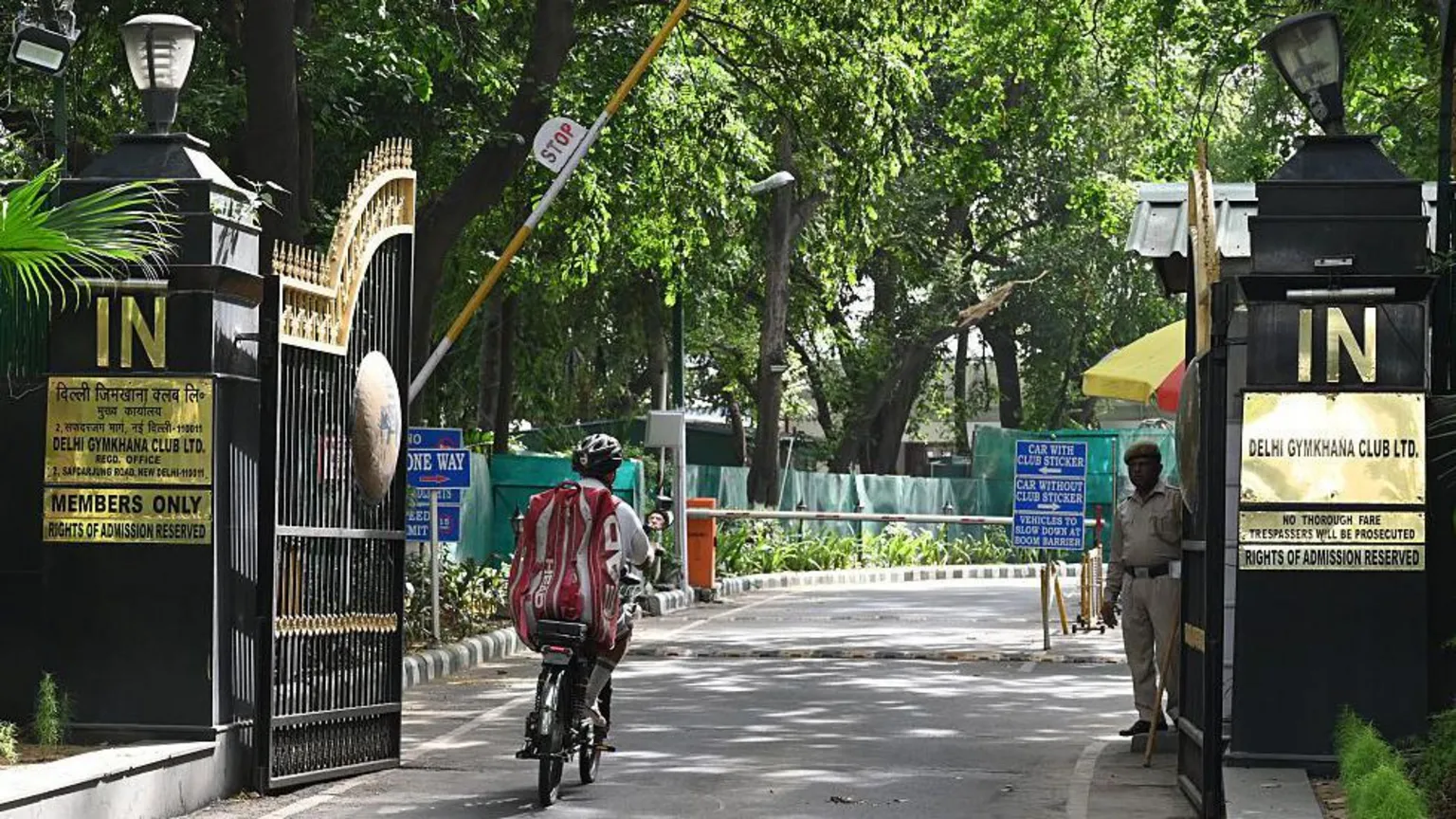  A uniformed guard stands at the gate of the Delhi Gymkhana Club as a cyclist zooms into the campus surrounded by trees. Plates on the gates and the wall mention 