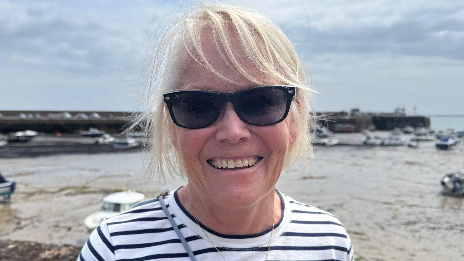 Elaine Davis pictured in Gorey Harbour. She is wearing a Breton-striped top, black sunglasses and has blonde hair. Behind her, the harbour is at low tide, and there are moored boats.