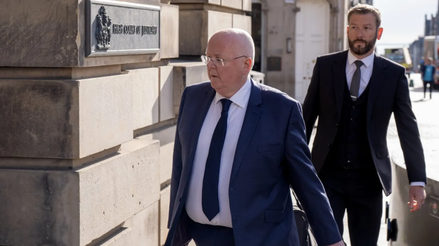 Two people in dark business suits walk along a sunlit pavement beside a stone building. The person in front is Peter Murrell and is wearing glasses and carrying a bag, passes a wall plaque reading “High Court of Justiciary.” A second suited person follows a few steps behind. The background shows a bright street scene with buildings and pedestrians out of focus.