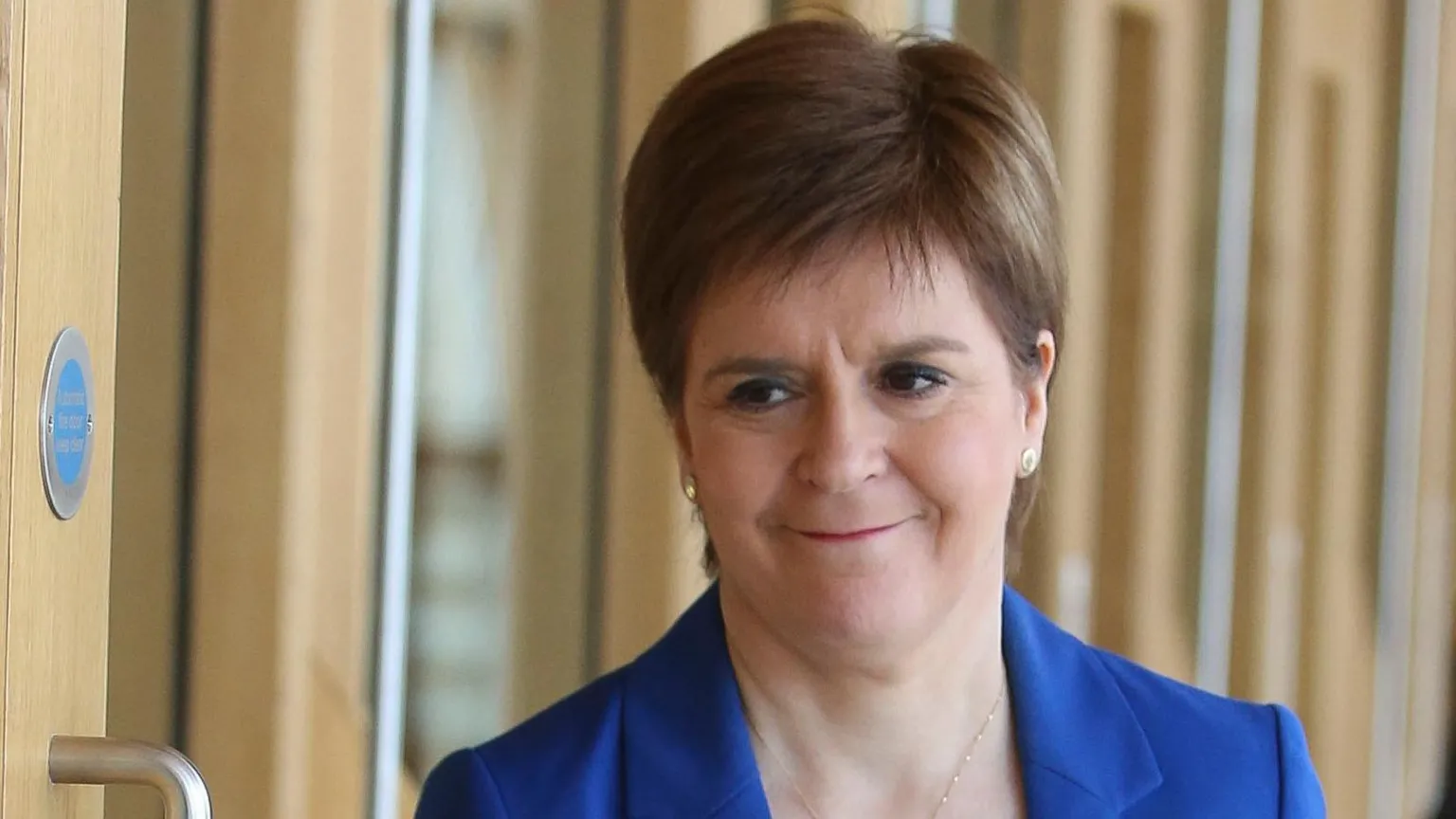 A close-up indoor photograph shows a Nicola Sturgeon who has short, neatly styled hair wearing a bright blue blazer over a light-coloured top and a round pendant necklace. She is standing near a wooden door with a visible metal handle and a small circular sign on the wall. The background consists of softly blurred wooden panels and glass, suggesting a modern interior space with natural light.